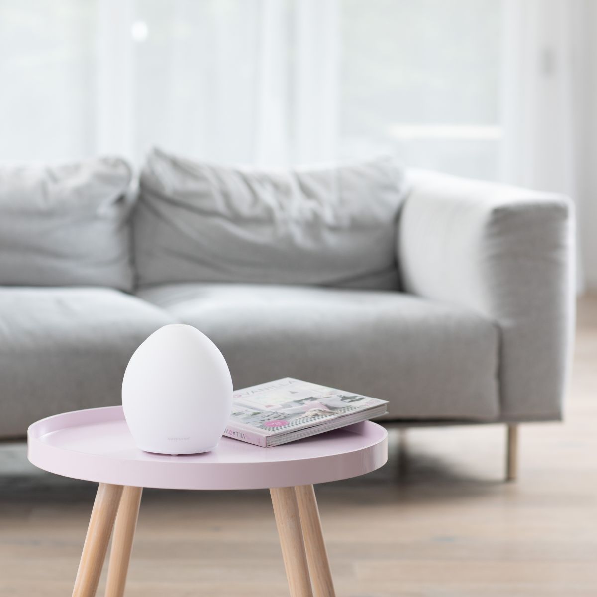 Ein moderner Aroma-Diffuser steht auf einem kleinen Tisch vor einem Sofa in einem hellen Wohnzimmer.